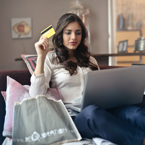 Eine Frau sitzt auf dem Sofa, neben ihr sind Shopping-Bags und sie hält eine Kreditkarte in der Hand. Auf ihrem Schoß ist ihr Laptop