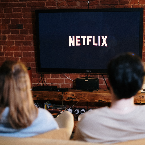 Ein Paar sitzt auf dem Sofa. Vor Ihnen hängt ein Fernseher an der Wand. Auf dem Bildschirm ist das "Netflix"-Logo zu sehen